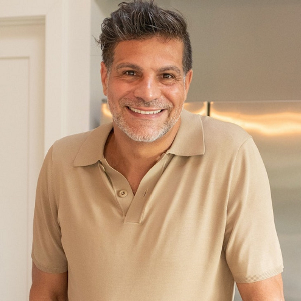 Smiling man with short, dark hair and a beard wearing a beige polo shirt, standing indoors near a refrigerator and a white door.