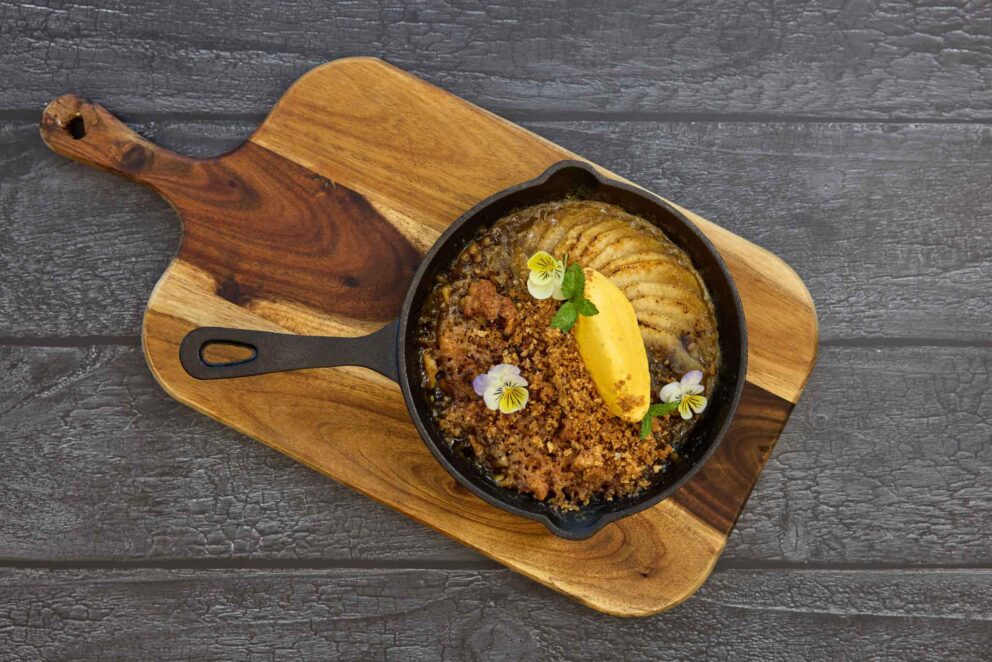 A cast iron skillet with baked fruit crumble, a scoop of yellow ice cream, edible flowers, and mint, served on a wooden board against a dark wood background.