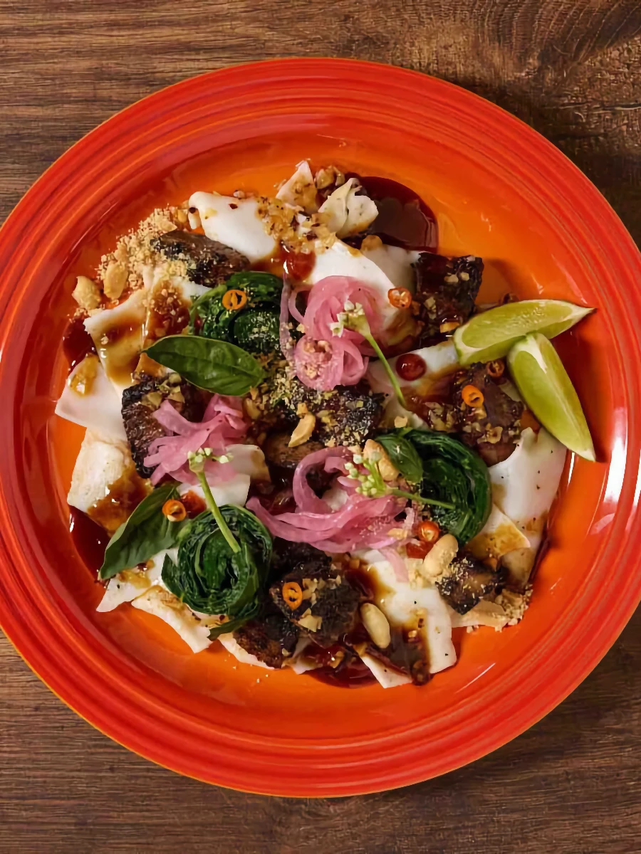 A bright orange plate holds rice noodles topped with pieces of meat, pickled onions, lime wedges, greens, crushed peanuts, and sliced chili peppers on a wooden surface.