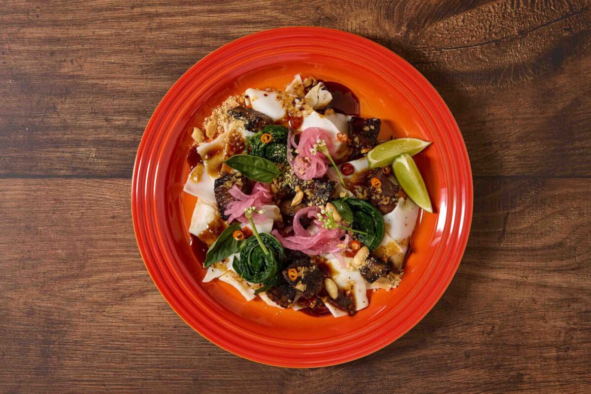 A red plate with a dish featuring pieces of meat or fish, leafy greens, pickled onions, lime wedges, and a creamy sauce, set on a wooden table.