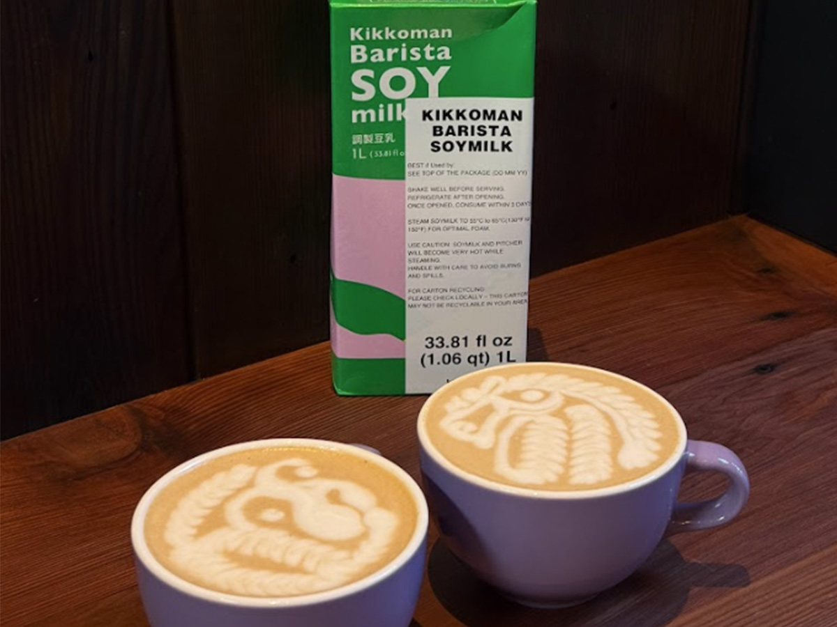 Two cups of latte with intricate latte art sit on a wooden table next to a carton of Kikkoman Barista Soy Milk.