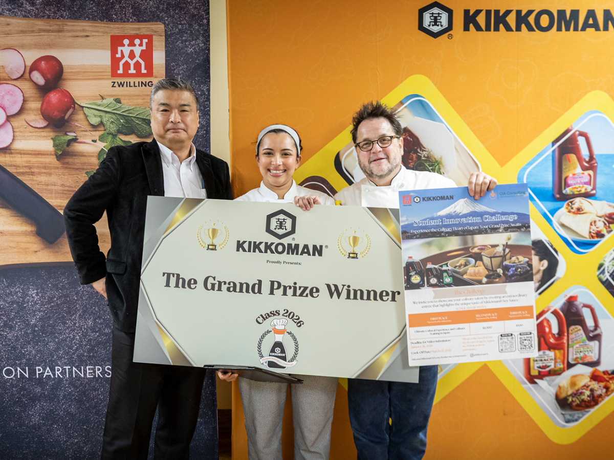 Three people stand together holding a large "Kikkoman Grand Prize Winner" sign and a poster, in front of orange Kikkoman and Zwilling branded banners.