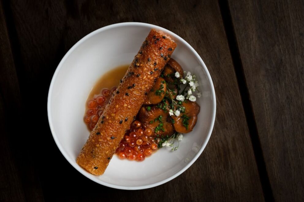 A white bowl with a crispy rolled cracker, salmon roe, mushrooms in sauce, and a garnish of herbs and small white flowers on a dark wooden surface.