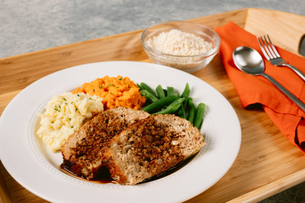 A plate with two slices of meatloaf, mashed potatoes, mashed sweet potatoes, and green beans on a wooden tray, with a small bowl of rice and silverware on an orange napkin.