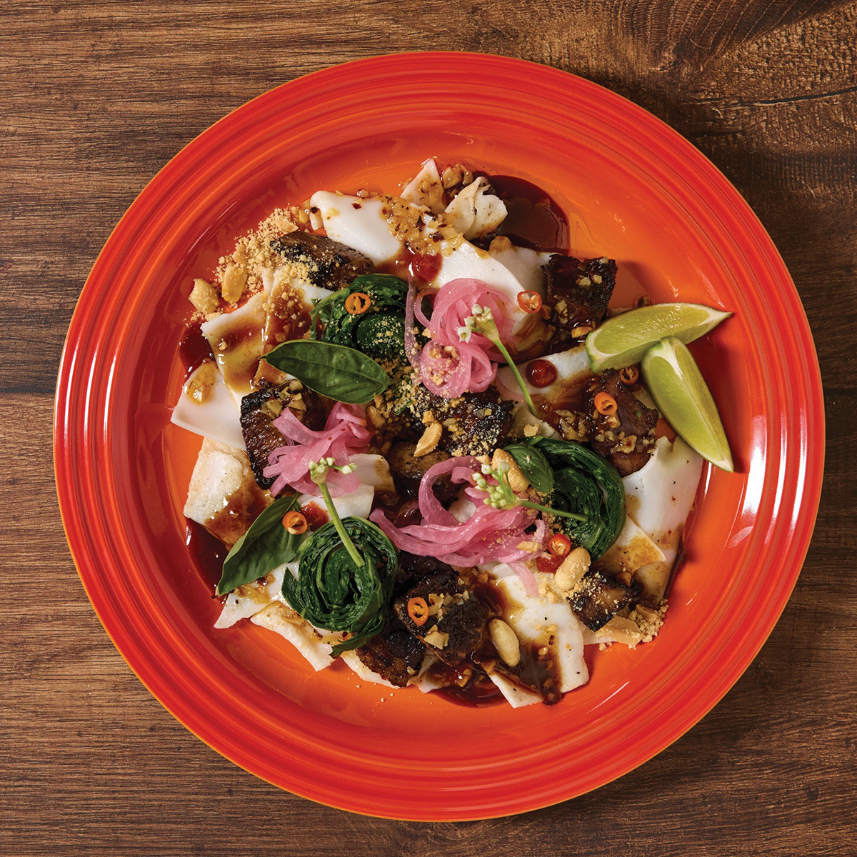 A red plate with rice noodles, grilled meat, pickled onions, leafy greens, chopped nuts, sliced chili, sauce, and lime wedges on a wooden table.