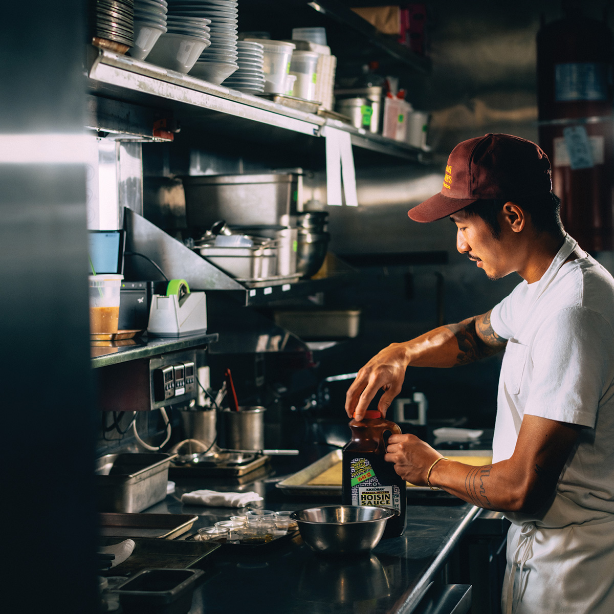 A person in a commercial kitchen opens a bottle of hoisin sauce and prepares ingredients at a counter.