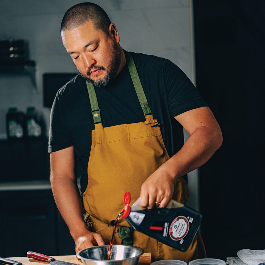 A man in a mustard apron pours soy sauce from a large container into a metal bowl in a kitchen setting.