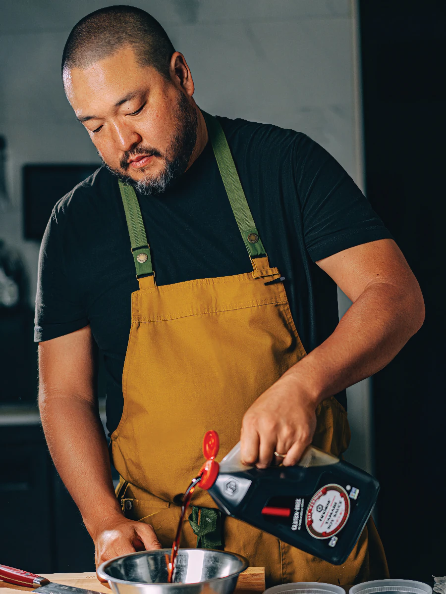 A man in a mustard apron pours soy sauce from a large container into a metal mixing bowl in a kitchen setting.