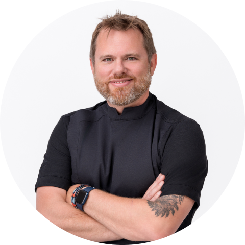 Man with light brown hair and beard, wearing a black shirt, standing with arms crossed and smiling, visible tattoo on left forearm, white background.