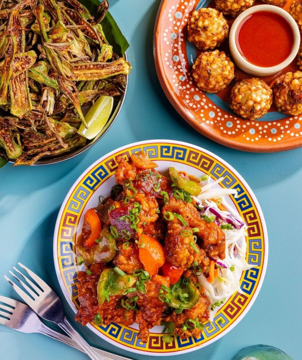 Three plates of food on a blue table: fried okra with lime, sauced vegetable stir-fry with slaw, and round fritters with a bowl of red dipping sauce. Fork and spoon on the side.