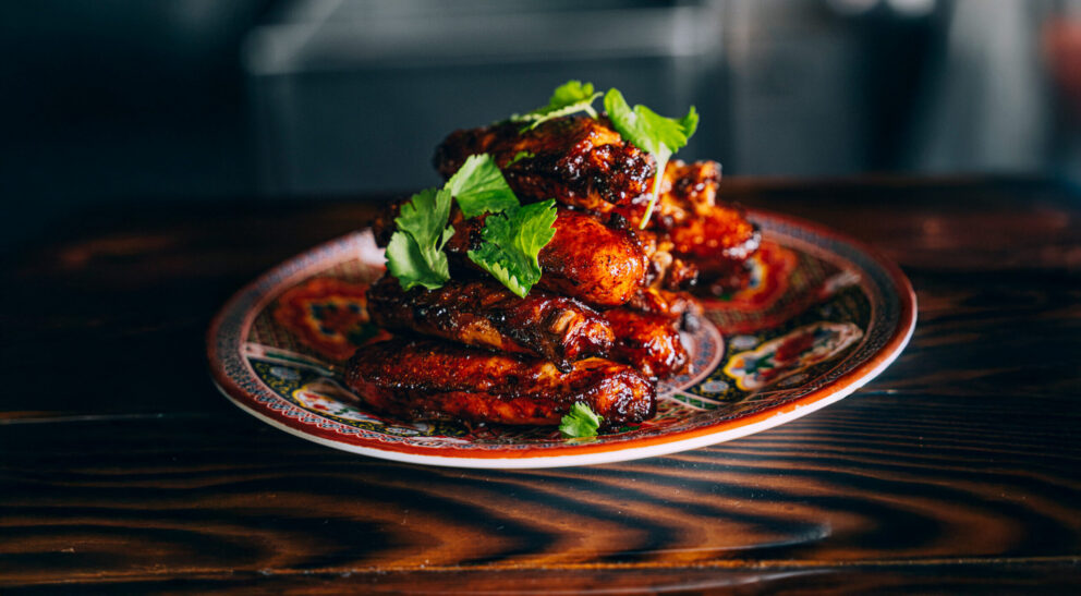 A stack of glazed chicken wings garnished with fresh cilantro sits on a patterned plate atop a dark wooden table.