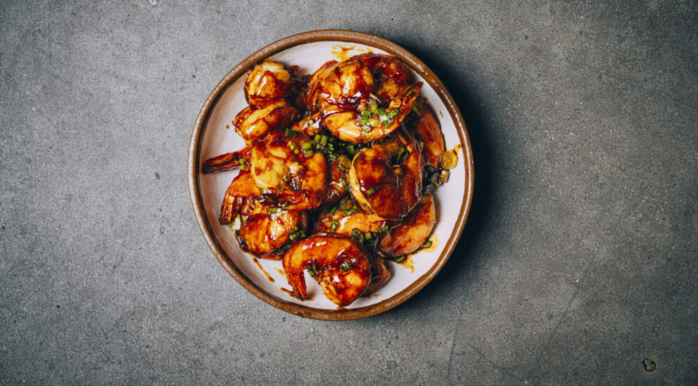 A plate of glazed shrimp garnished with chopped green herbs sits on a gray textured surface.