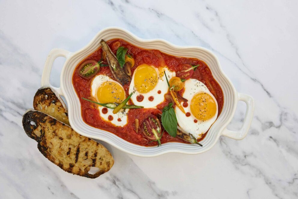 Baked eggs in tomato sauce garnished with herbs and vegetables, served in a white dish with two slices of grilled bread on the side.