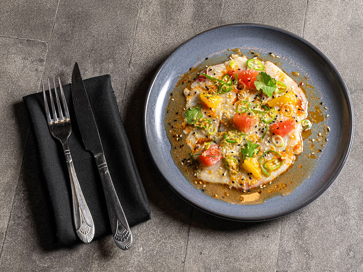 A blue plate with ceviche garnished with cilantro, sliced peppers, and citrus segments, next to a black napkin with a fork and knife on a gray stone surface.