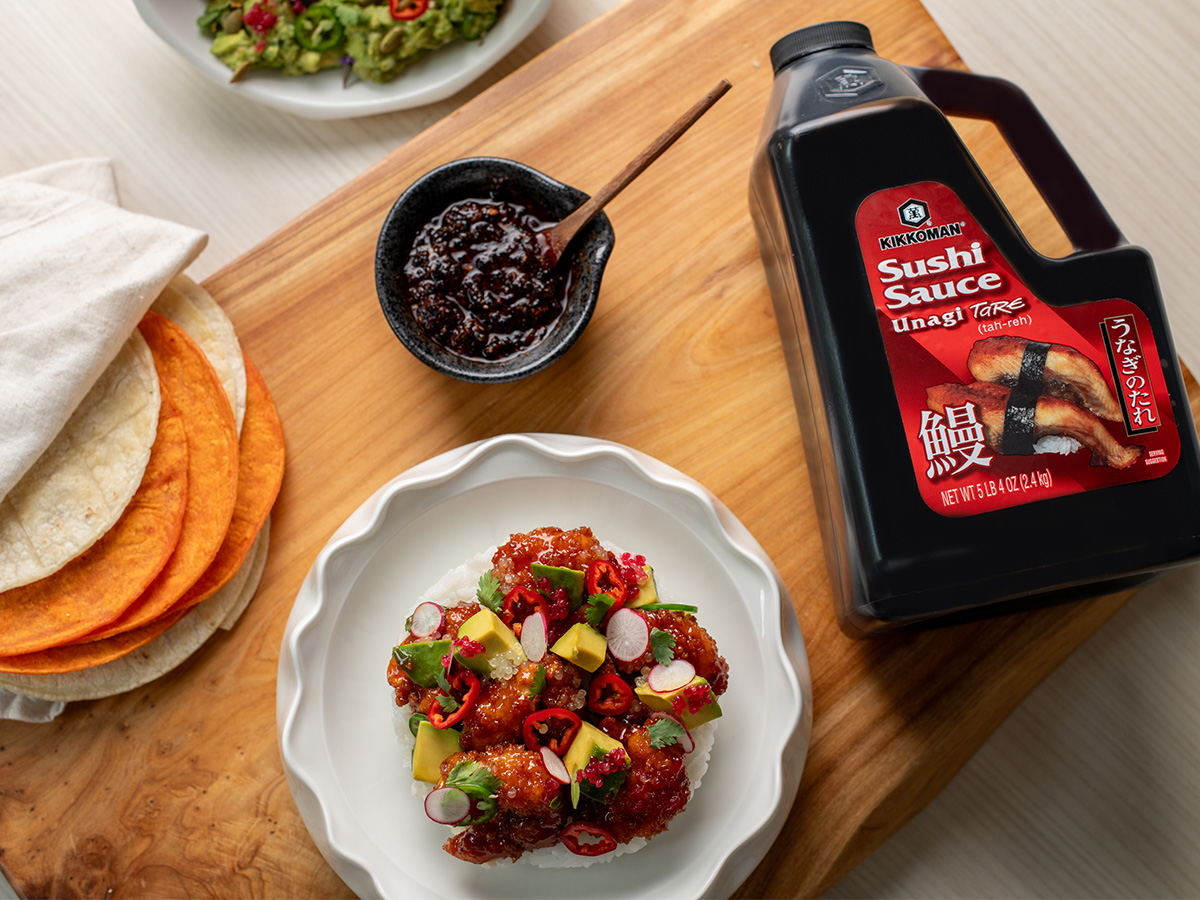 A white plate of fried chicken pieces topped with avocado, radish, and chili sits beside tortillas, a bowl of dark sauce, and a large bottle of Kikkoman Sushi Sauce on a wooden board.