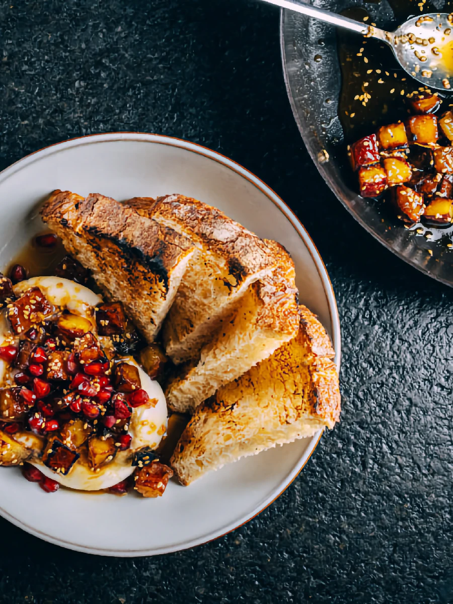 A plate with slices of toasted bread, creamy cheese, and a topping of diced fruit and seeds, next to a bowl with more fruit mixture and a spoon.