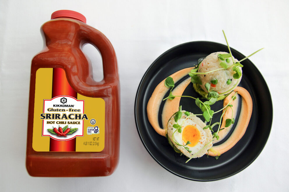 A large bottle of Kikkoman gluten-free Sriracha hot chili sauce sits next to a black plate with a gourmet dish garnished with sauce and herbs.