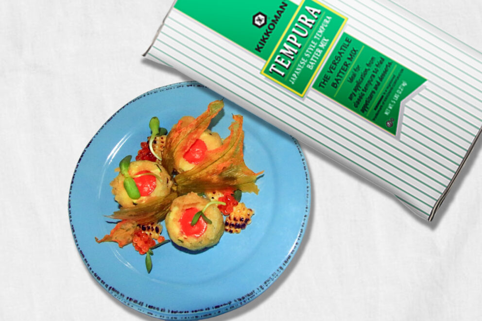 A blue plate with vegetable tempura garnished with corn and sauce sits next to a box of Kikkoman Tempura Batter Mix on a white background.
