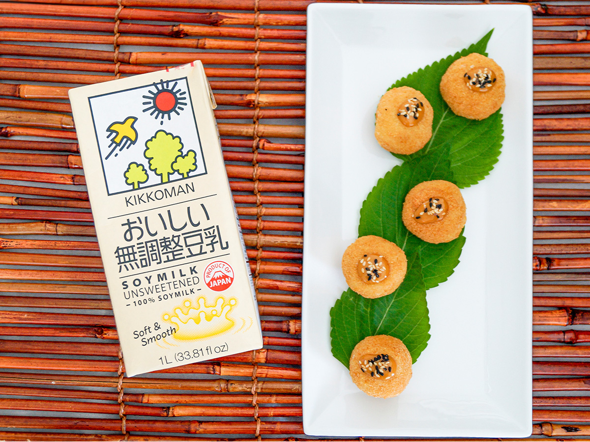 A carton of Kikkoman unsweetened soy milk sits beside a plate with five round cookies garnished with seeds, on green leaves, atop a bamboo mat.