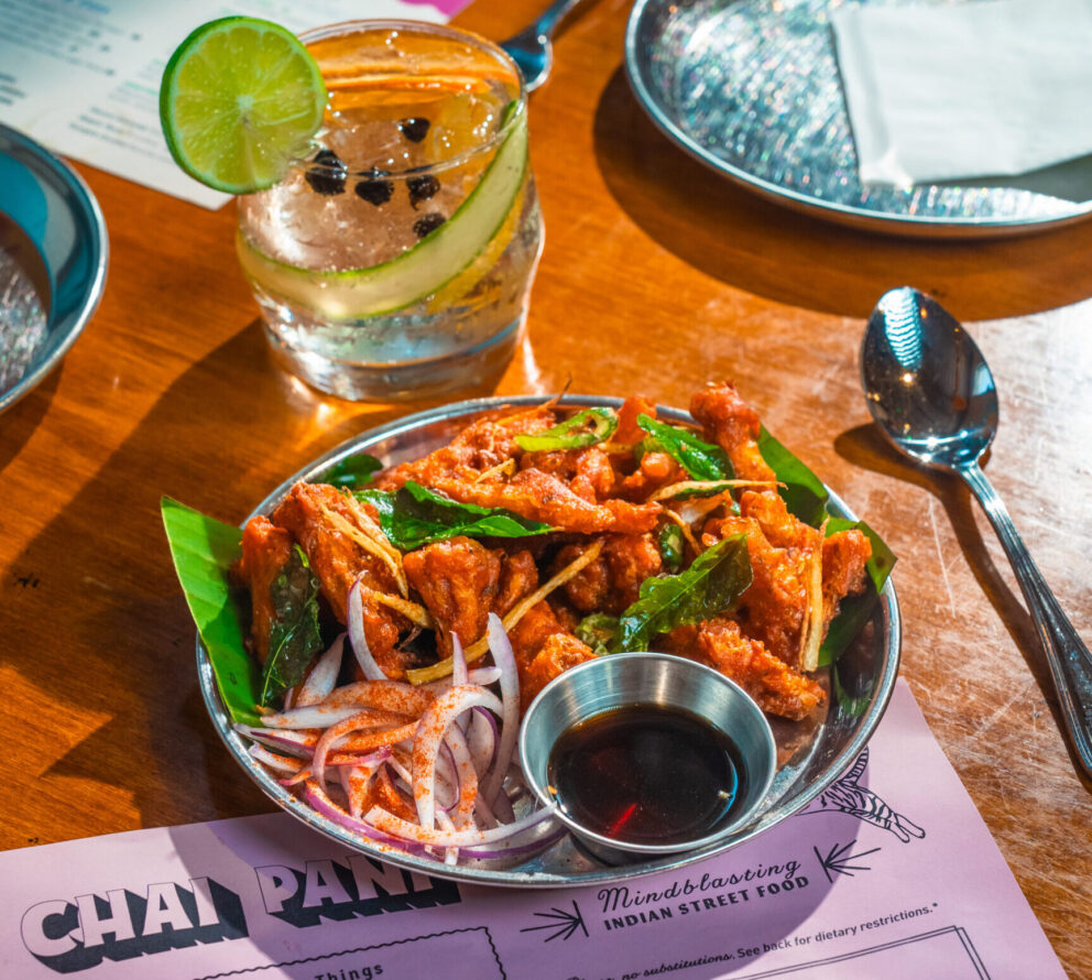 A plate of fried chicken garnished with curry leaves and sliced onions, served with dipping sauce, next to a cocktail with lime, cucumber, and black peppercorns.