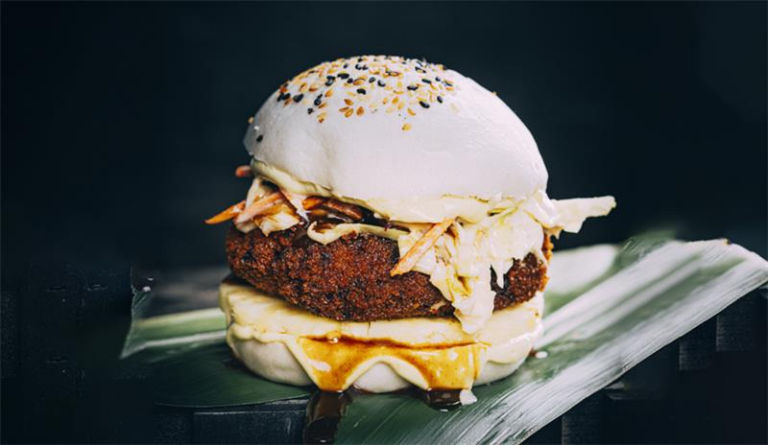 A sandwich with a breaded patty, shredded cabbage, sauce, and a white bun topped with sesame seeds, presented on a green leaf.