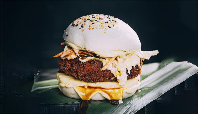 A sandwich with a breaded patty, shredded cabbage, sauce, and a white bun topped with sesame seeds, presented on a green leaf.