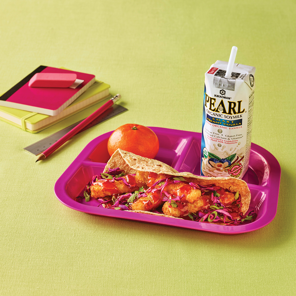 A pink tray with two breaded cauliflower tacos, an orange, a carton of soymilk, and a stack of notebooks with a ruler, eraser, and pencil on a green surface.