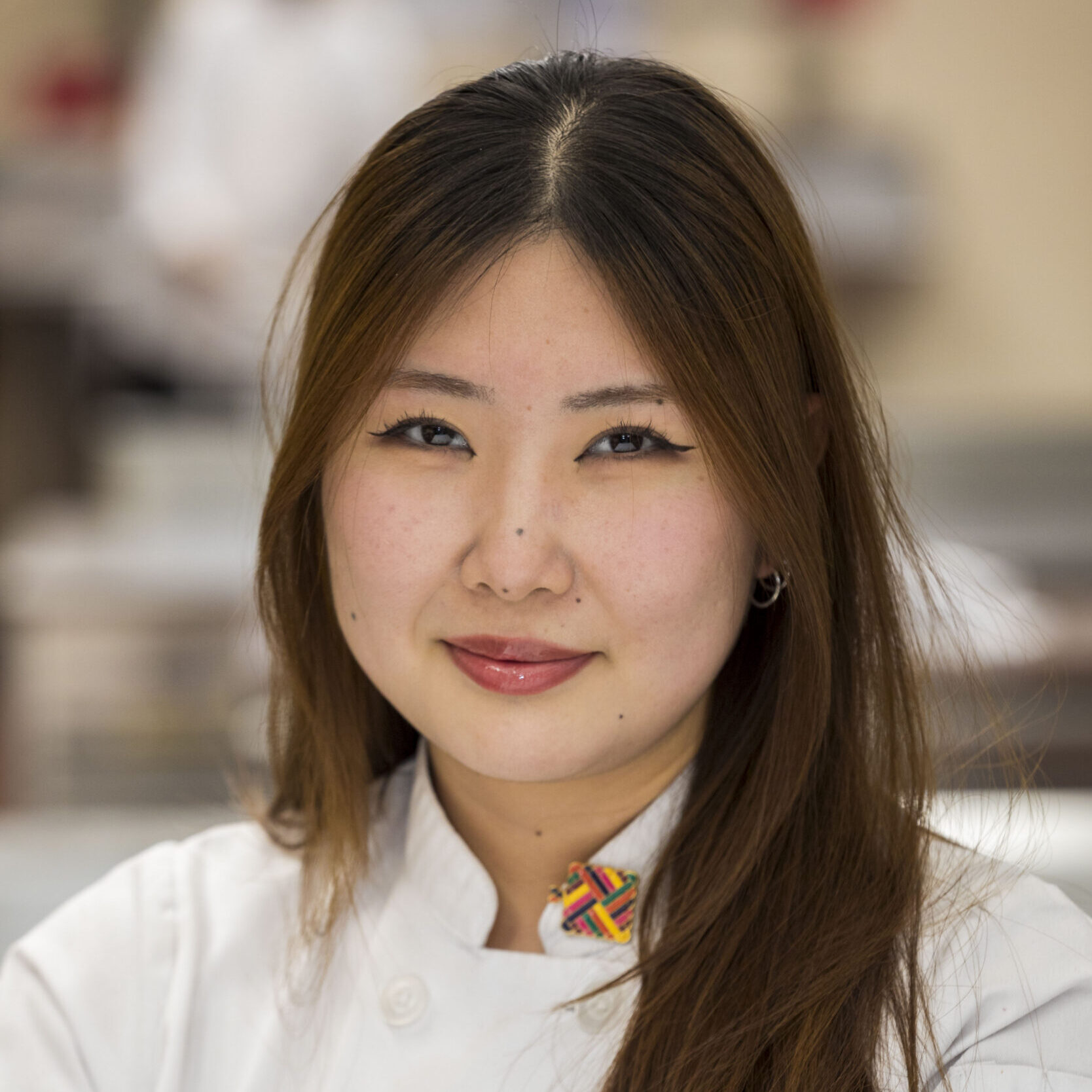 A woman with long brown hair wearing a white chef's coat and a colorful pin, standing indoors with a neutral expression.