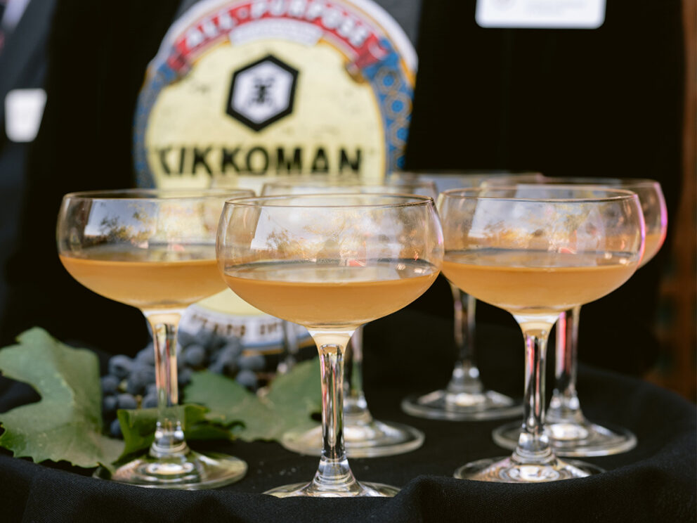 Six coupe glasses filled with a light amber beverage are arranged on a black tablecloth, with a person wearing a Kikkoman shirt in the background.