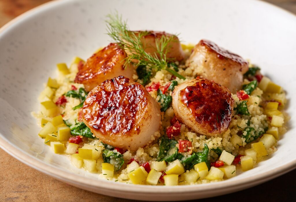 Four seared scallops sit on a bed of couscous mixed with diced vegetables and greens, garnished with a sprig of fresh dill in a shallow white bowl.