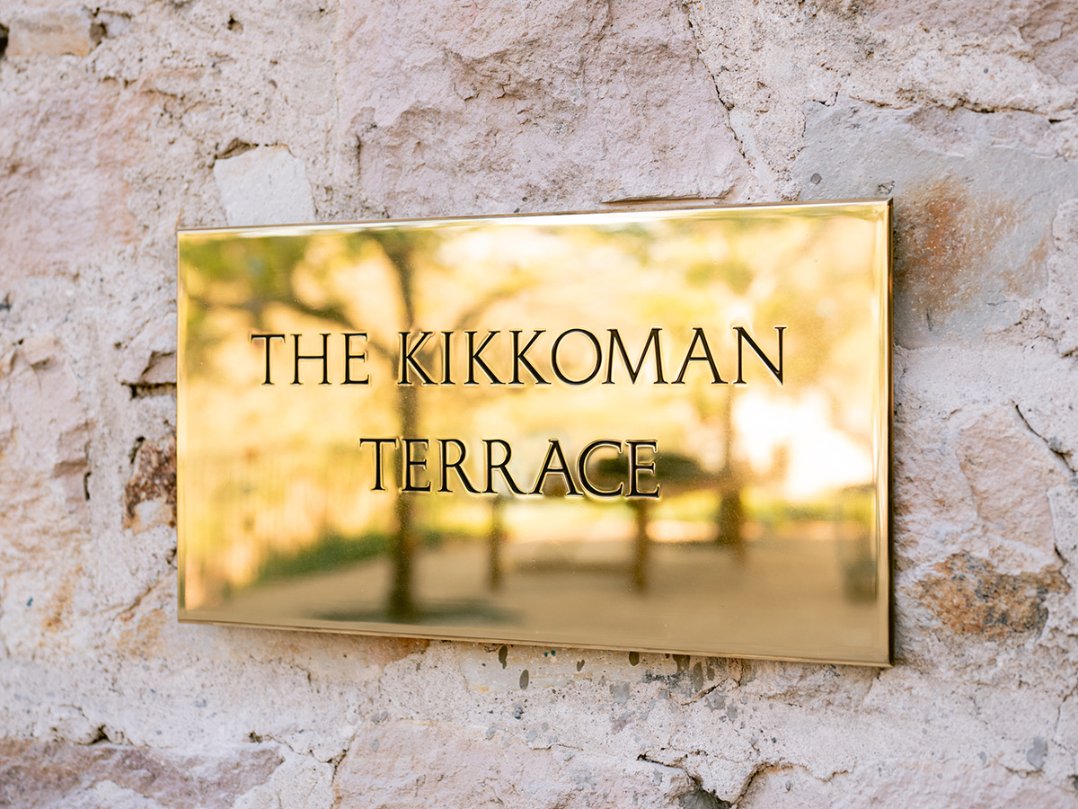 A gold plaque mounted on a stone wall reads “THE KIKKOMAN TERRACE” with a blurred reflection of trees and outdoor surroundings.