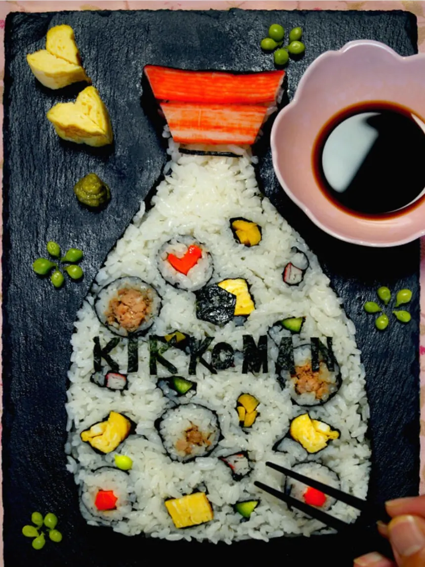 Sushi arranged in the shape of a soy sauce bottle with "Kikkoman" spelled in nori, served on a slate plate with soy sauce, tamagoyaki pieces, and green garnishes.