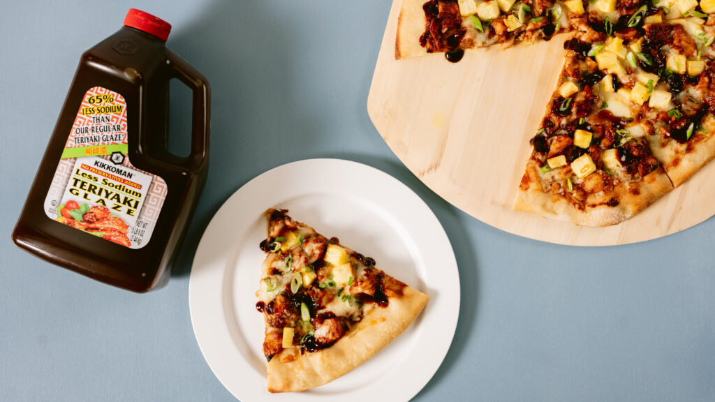 A slice of pizza on a white plate next to a bottle of Kikkoman teriyaki sauce; the rest of the pizza is on a wooden board.