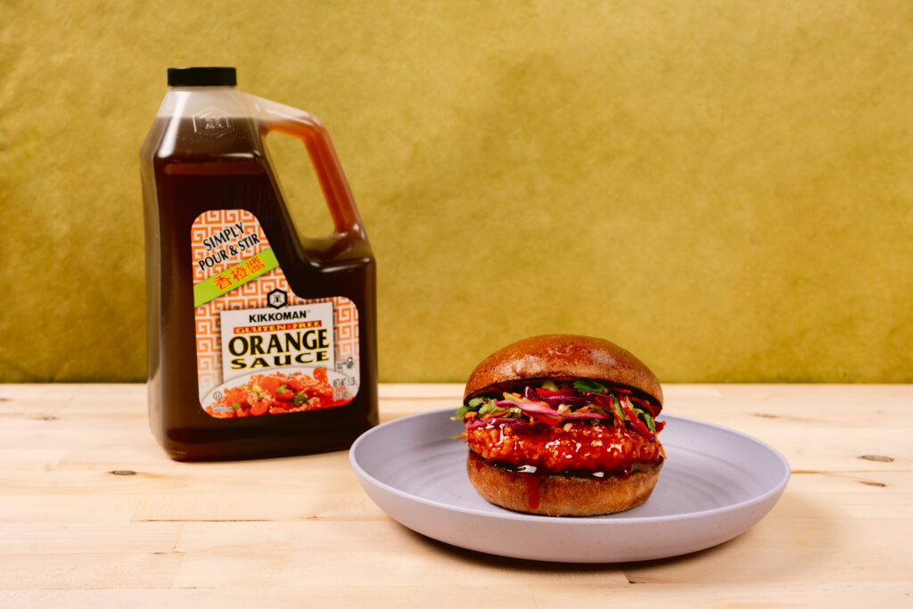 A bottle of Kikkoman Orange Sauce stands next to a chicken sandwich topped with greens and sauce on a blue plate, set on a light wooden surface with a yellow background.