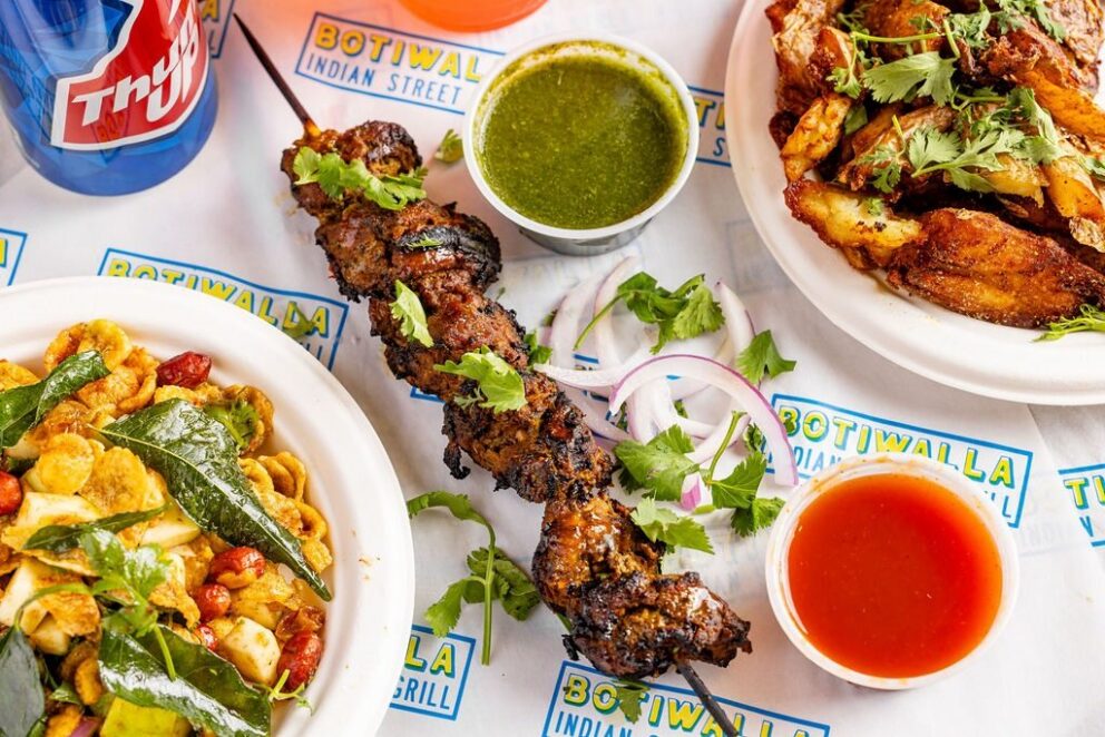 A skewer of grilled meat, potato wedges, green and red sauces, and a dish with curry leaves and snacks are arranged on a table with a drink cup and Botiwalla branding.