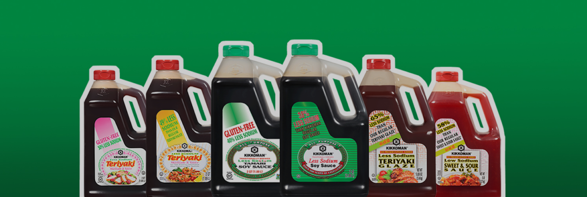A row of seven large Kikkoman sauce bottles, including soy sauce, teriyaki, sweet and sour, teriyaki glaze, and gluten-free varieties, against a green background.