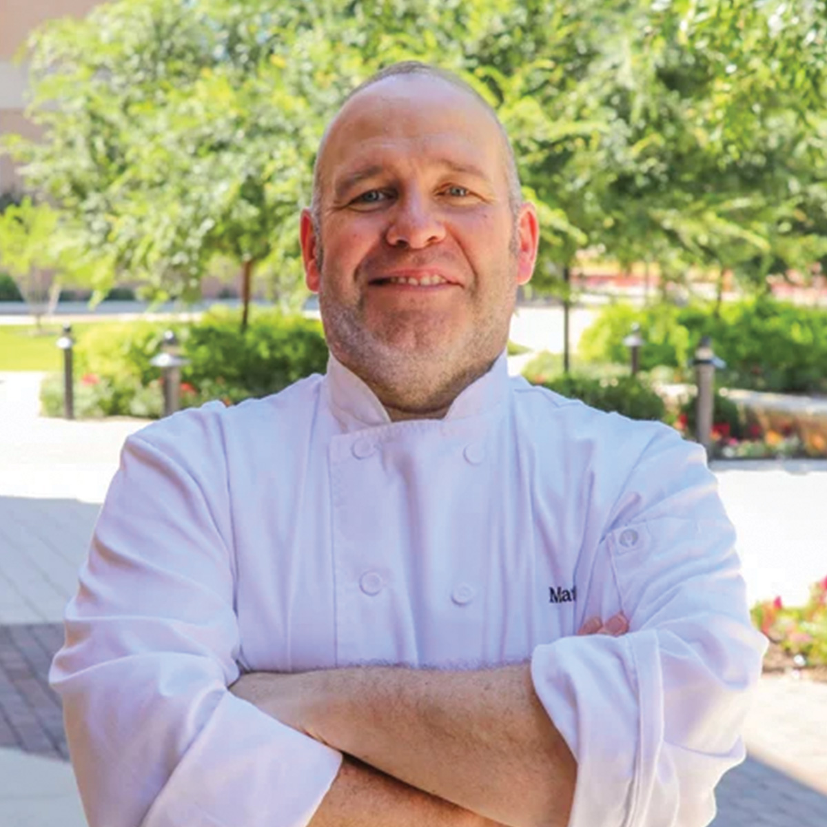 A chef wearing a white double-breasted coat stands outside with arms crossed, smiling, with greenery and a walkway in the background.