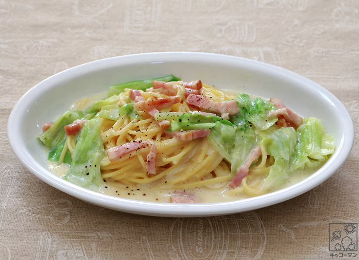 A white oval plate with creamy pasta topped with sliced bacon, cabbage, and black pepper, set on a beige tablecloth.