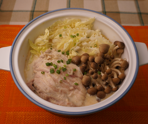 A ceramic bowl contains cooked chicken, cabbage, brown beech mushrooms, and chopped chives in a creamy broth, set on an orange placemat.