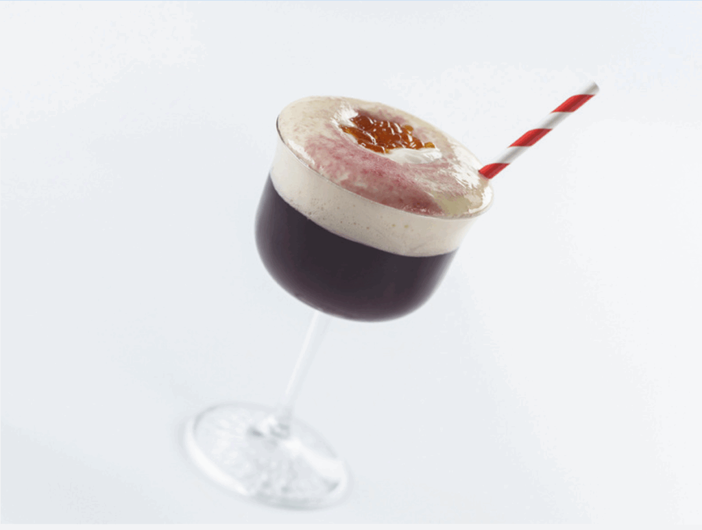 A dark cocktail in a stemmed glass topped with thick foam, a caramelized sugar garnish, and a red-and-white striped paper straw.