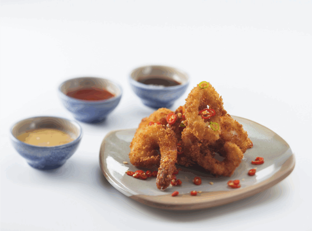 A plate of fried breaded shrimp and calamari garnished with sliced red chilies, with three dipping sauces in small blue bowls in the background.