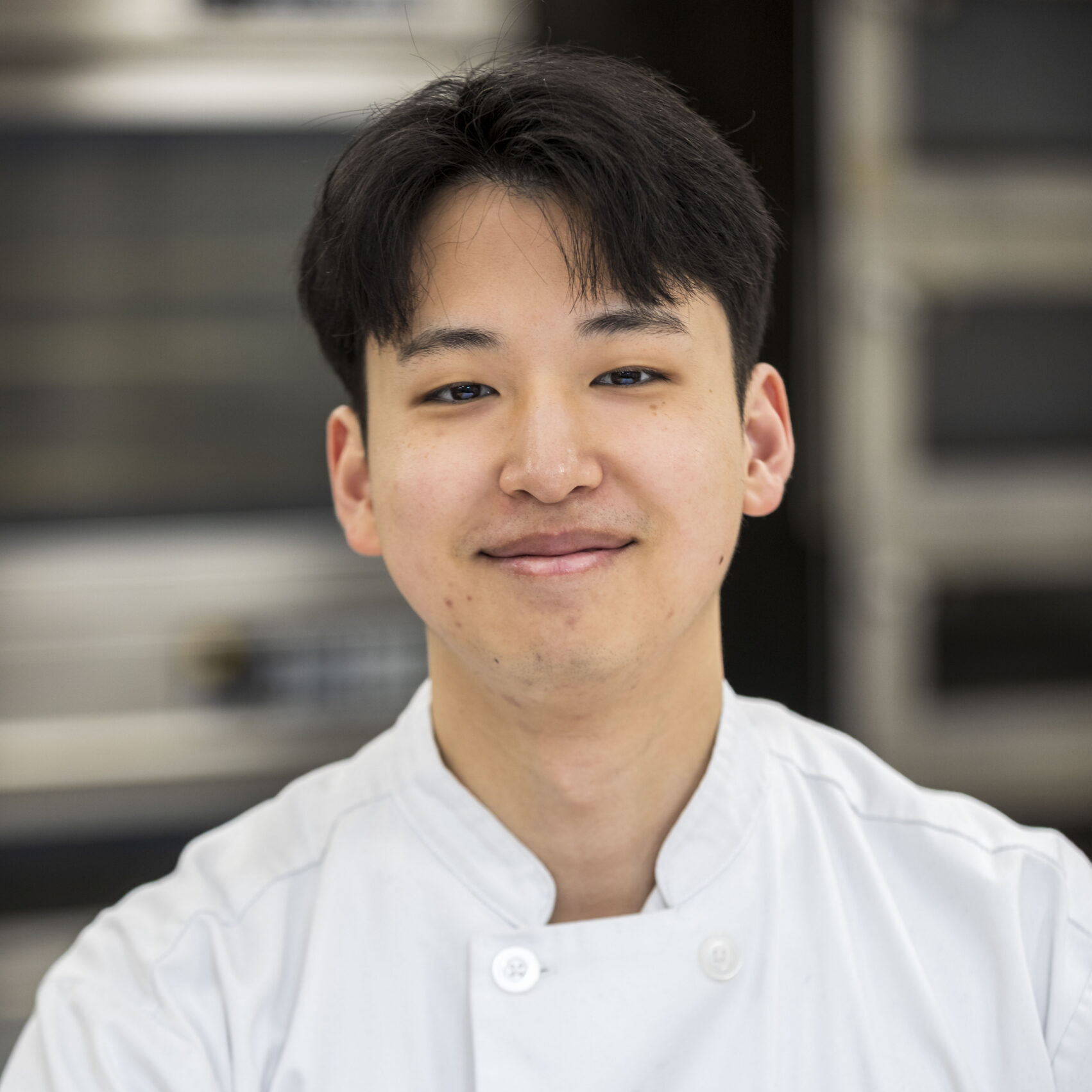 A man wearing a white chef's coat is smiling slightly while looking at the camera. The background is blurred kitchen equipment.