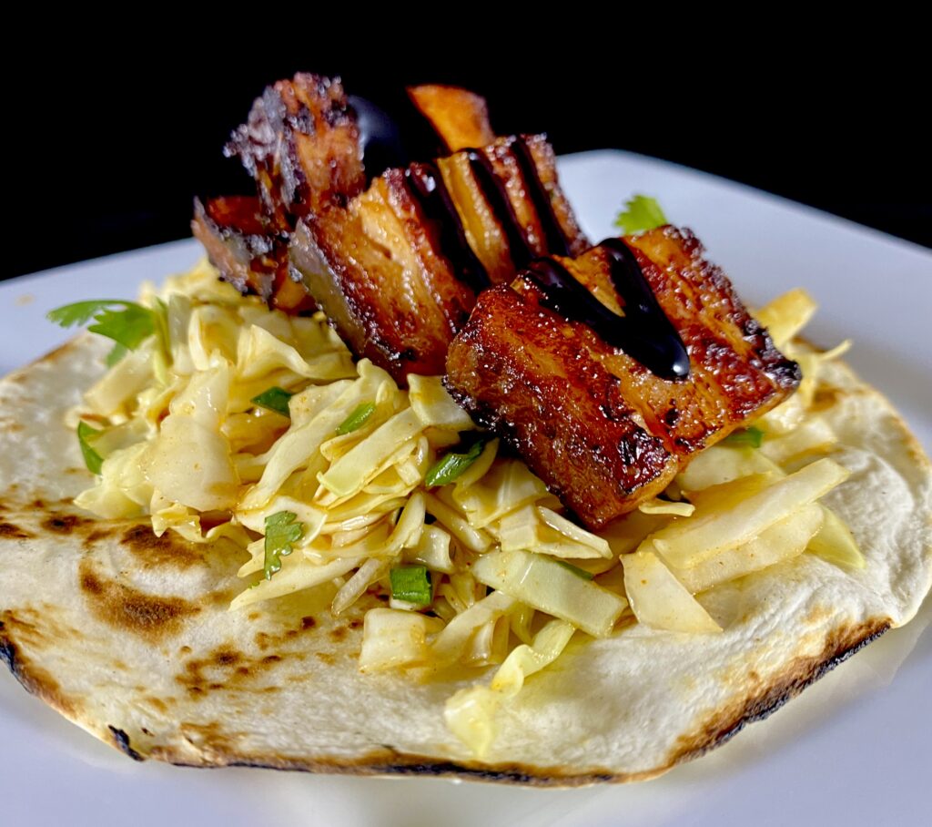 A flour tortilla topped with shredded cabbage, cilantro, and thick pieces of glazed pork belly, drizzled with a dark sauce, on a white plate.