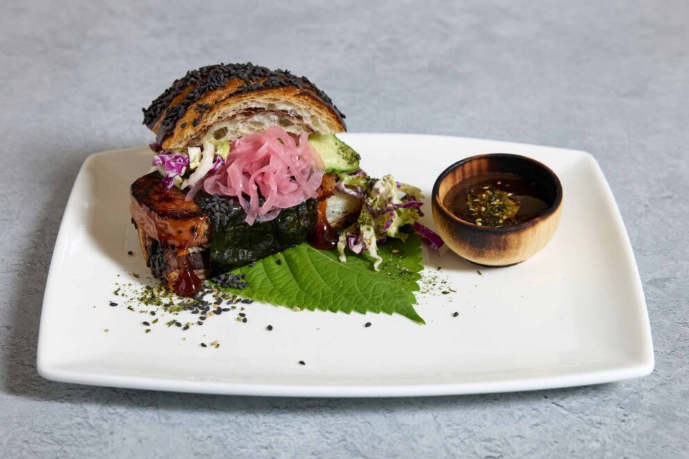 A plated dish with sliced bread, glazed meat wrapped in seaweed, pickled onions, slaw, a green leaf, black sesame seeds, and a small wooden bowl of dipping sauce on a white square plate.