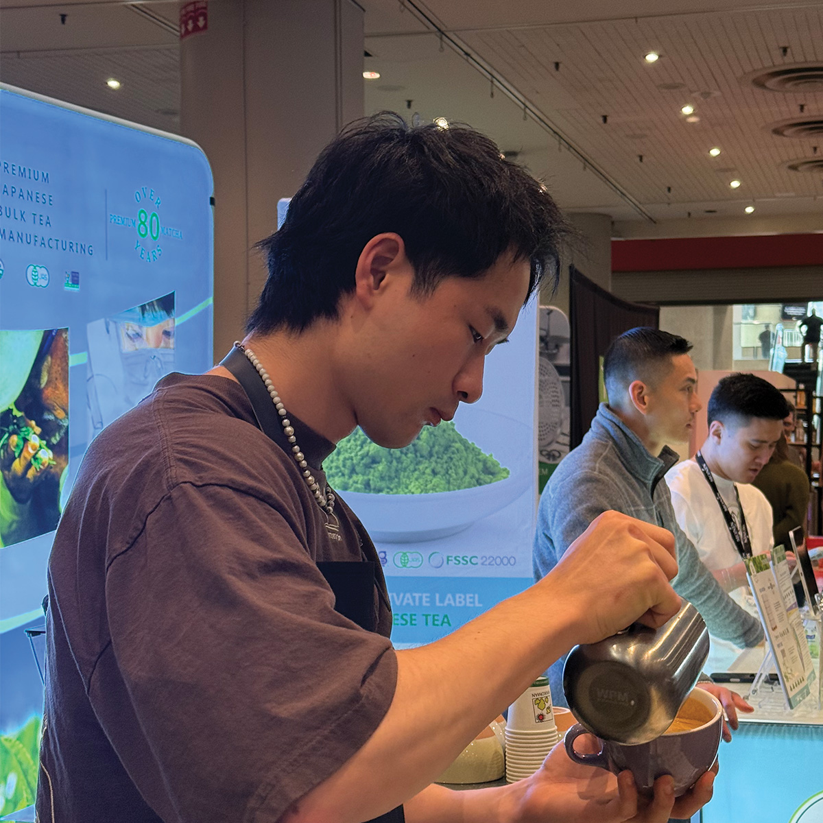 A person pours liquid from a metal pitcher into a cup at a booth, with two other people standing nearby and promotional banners in the background.