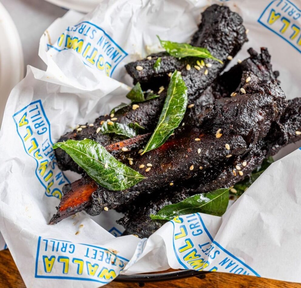 A basket lined with branded paper contains several pieces of charred meat garnished with green leaves and sesame seeds.