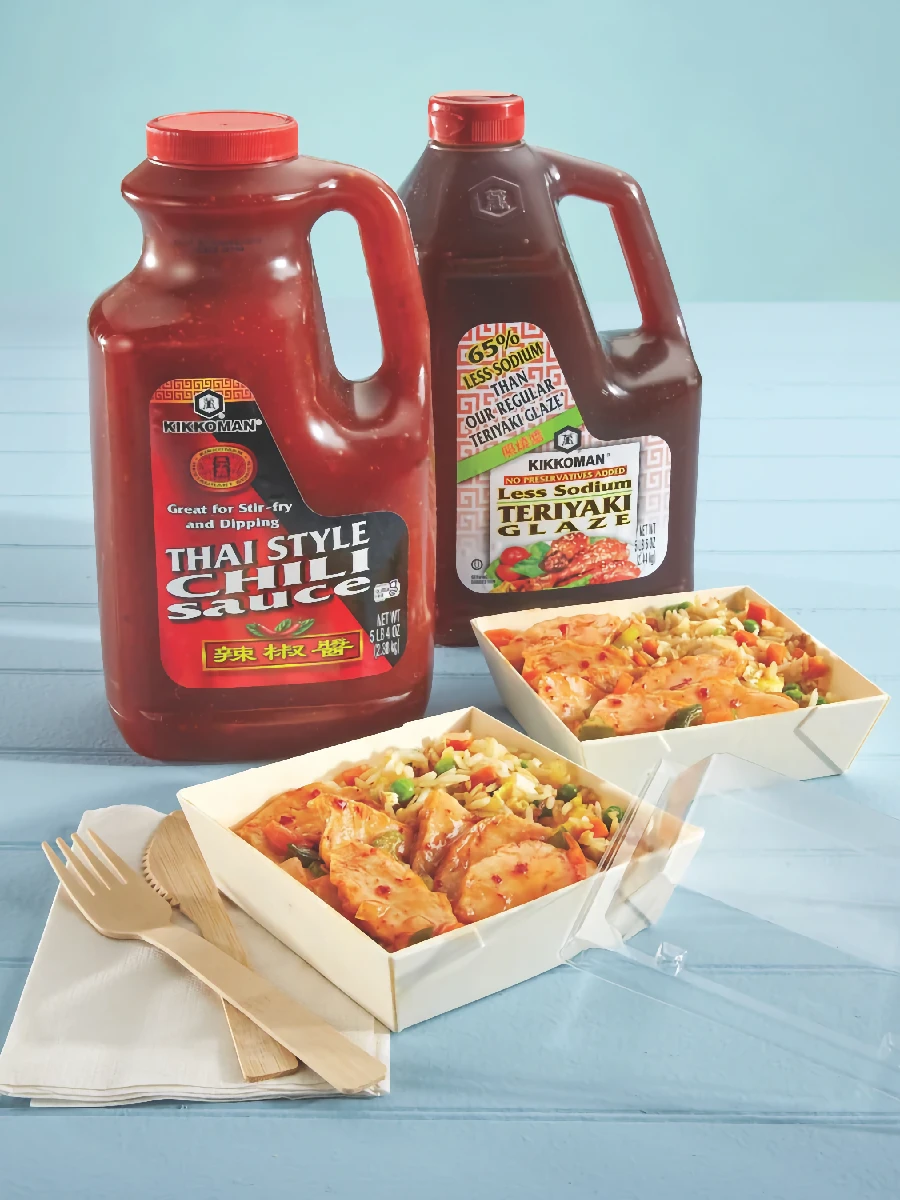 Two large bottles of Kikkoman sauces and two takeout containers of stir-fry with chicken and vegetables, placed on a blue surface with wooden utensils.