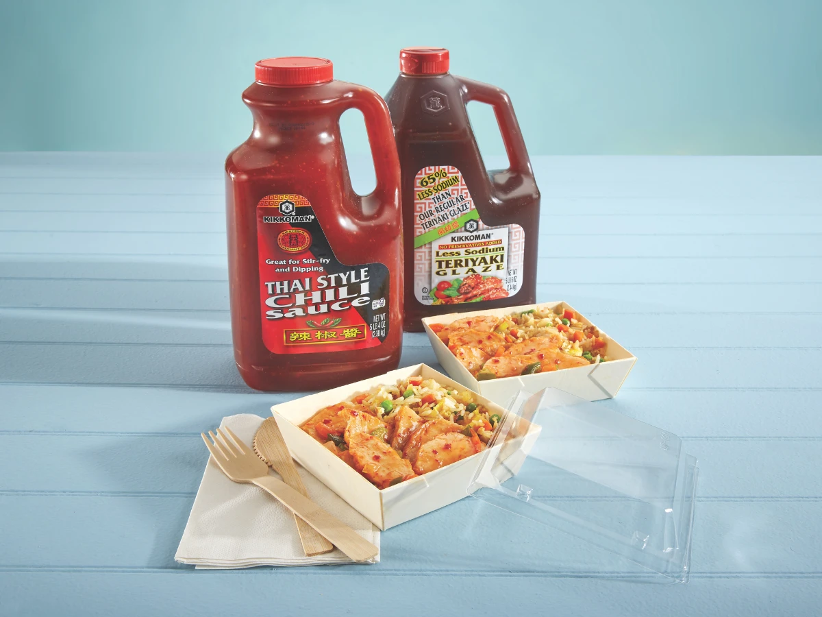 Two large bottles of Thai-style chili sauce and teriyaki sauce are placed behind two takeout containers of stir-fried noodles with vegetables, with wooden cutlery and napkins on the side.
