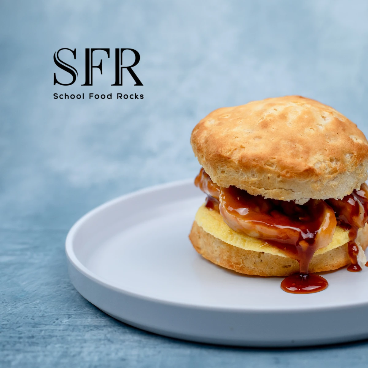 A biscuit sandwich with egg and barbecue sauce sits on a white plate, with the "School Food Rocks" logo in the background.