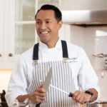 A chef in a white coat and striped apron is smiling while sharpening a knife with a honing rod in a kitchen.